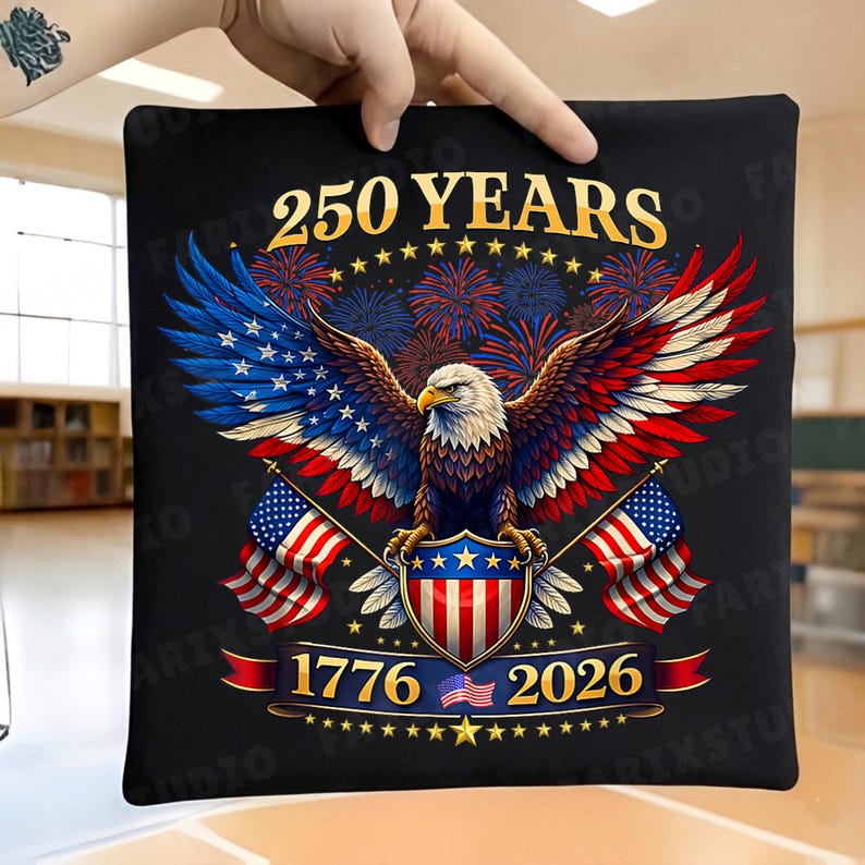 May include: Black square pillow featuring a patriotic design with an eagle, American flags, and fireworks. The text reads "250 YEARS 1776-2026". The eagle has blue, red, and white wings. The pillow is held by a hand.