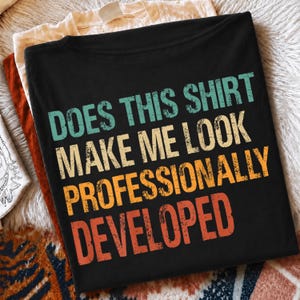 Op de afbeelding: Een zwart t-shirt met de tekst "DOES THIS SHIRT MAKE ME LOOK PROFESSIONALLY DEVELOPED" in een verweerd, meerkleurig lettertype. De woorden zijn in rijen gestapeld met groenblauwe, gele en oranje tinten, wat een vintage grafisch ontwerp creëert.
