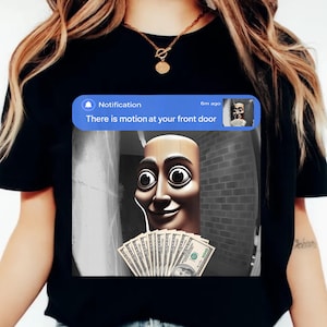 May include: Black t-shirt featuring a humorous graphic. The graphic shows a notification bubble with the text "There is motion at your front door." Below the notification is a cartoon face holding a fan of hundred-dollar bills.