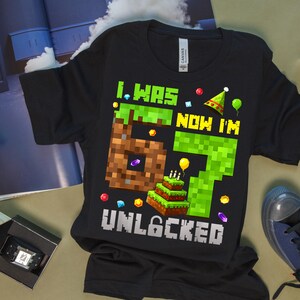 May include: Black t-shirt with a pixelated design. The design includes the text "I WAS 6 NOW I'M 7 UNLOCKED" with a pixelated number 6, a pixelated number 7, a birthday cake, and other pixelated graphics.