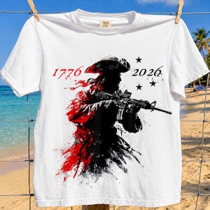 以下が含まれることがあります： 白いTシャツ。シルエットでライフルを構える人物のグラフィック、赤と黒のペイントスプラッター効果。数字の「1776」と「2026」がプリントされ、黒い星が3つ描かれています。
