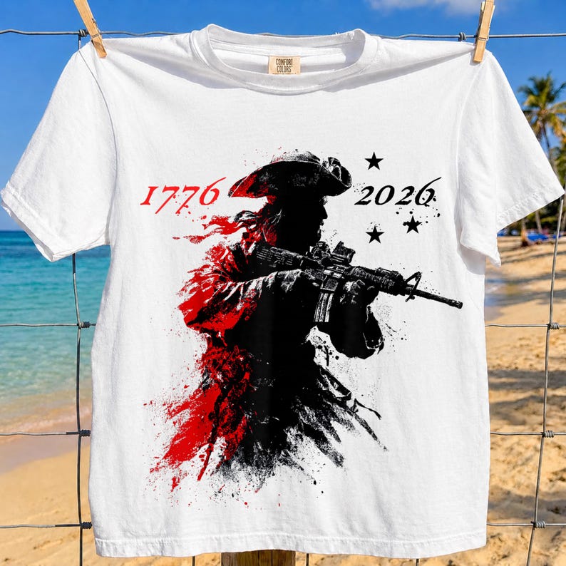 以下が含まれることがあります： 白いTシャツ。シルエットでライフルを構える人物のグラフィック、赤と黒のペイントスプラッター効果。数字の「1776」と「2026」がプリントされ、黒い星が3つ描かれています。