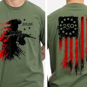以下が含まれることがあります： オリーブグリーンのTシャツ。ライフルを持った兵士のグラフィック、数字の「1776」と「2026」、星が描かれています。背中には、数字「250」が入った、使い古されたアメリカ国旗のデザインが施されています。