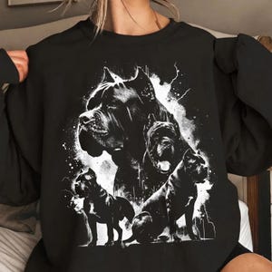 May include: A black crewneck sweatshirt featuring a high-contrast white graphic print of three Cane Corso dogs. The design includes one large portrait of a dog head at the top, with two smaller full-body dog figures positioned below.