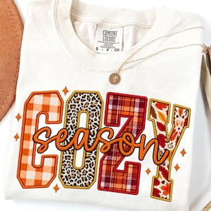 May include: A cream-coloured short-sleeve t-shirt featuring the word COZY in large block letters with varied patterns including orange plaid, leopard print, red plaid, and autumn leaves, with the word season in orange cursive script.
