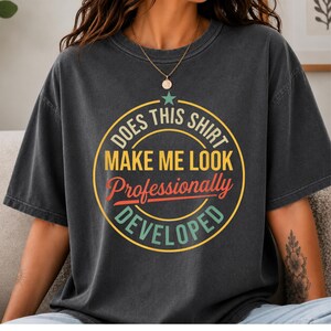 May include: A charcoal grey short-sleeve t-shirt featuring a circular graphic with a star at the top. The text reads "DOES THIS SHIRT MAKE ME LOOK Professionally DEVELOPED" in yellow, red, and teal colours, centred on the chest of the garment.
