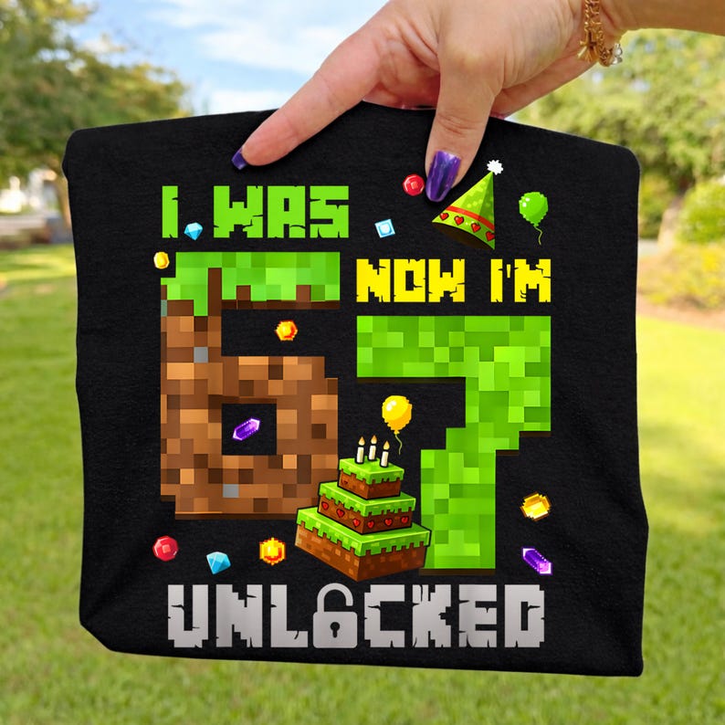 May include: Black t-shirt with a pixelated design. The text reads "I WAS 6, NOW I'M 7 UNLOCKED" in green and yellow. The design includes a pixelated number 6, a birthday cake, and colorful pixelated decorations.