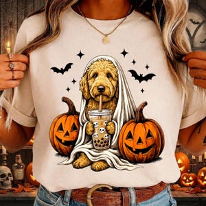 May include: A cream-colored t-shirt featuring a graphic of a golden-colored dog draped in a white sheet like a ghost, holding a bubble tea cup with skull designs. The dog is flanked by two orange jack-o'-lanterns and surrounded by black bats.