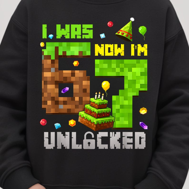 May include: Black sweatshirt with a colorful graphic design. The design features the text "I WAS NOW I'M 67 UNLOCKED" in pixelated green and brown blocks, with a birthday cake and party hat.