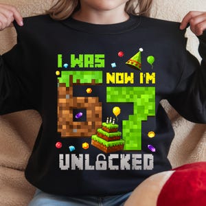 May include: Black sweatshirt with a colorful graphic design. The design features the text "I WAS 6 NOW I'M 7 UNLOCKED" in pixelated font, along with a pixelated cake, balloons, and party hat. The number 6 is brown and the number 7 is green.