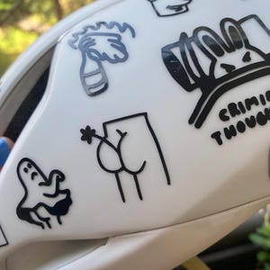 Könnte beinhalten: Ein weißer Helm, der mit verschiedenen schwarzen Vinylaufklebern verziert ist. Die Motive umfassen einen Geist, eine brennende Flasche, eine Zeichentrickfigur, einen Stern, einen Hasen mit dem Text CRIMINAL THOUGHTS und eine Strichzeichnung von Gesäß mit einer Blume.