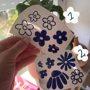 Könnte beinhalten: Zwei weiße Klebebögen mit dunkelblauen Blumenaufklebern. Der obere Bogen zeigt fünf einfache fünfblättrige Blumen in Umriss- und Volltonstilen. Der untere Bogen enthält sechs verschiedene Blumendesigns, darunter ein Gänseblümchen.