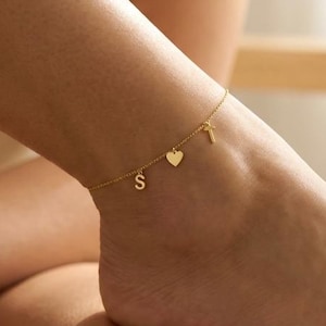 Könnte beinhalten: Goldfarbenes Fußkettchen mit drei Anhängern: dem Buchstaben "S", einem Herz und einem kleinen Schlüssel. Das Fußkettchen wird am Knöchel getragen, Fuß und Unterschenkel sind sichtbar. Der Hintergrund ist unscharf.