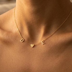 May include: A gold-colored necklace with the letters "D", "E", and a heart charm. The necklace is worn around the neck and rests on a person's chest. The person is wearing a black top.