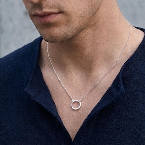 May include: A silver-toned circular pendant suspended on a delicate matching chain. The pendant features a simple, smooth ring design. The necklace is worn against a dark navy blue textured shirt, highlighting the metallic finish of the jewelry.