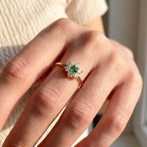 May include: A gold ring featuring a flower-shaped design. The ring has a central green gemstone surrounded by smaller white gemstones. The ring is on a finger, with a cream-colored top in the background.