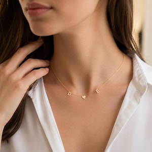 May include: A delicate gold-toned necklace featuring a thin chain with three small charms: a capital letter D, a centered heart shape, and a capital letter E. The necklace rests against the neck, showcasing a minimalist design with polished metal.