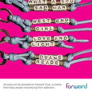 May include: Keyrings featuring white letter beads spelling phrases such as "WHAT A SAD MAN" and "WEST END GIRL." The keyrings have blue cords and gold-coloured accents, set against a bright pink background. Each keyring has a gold-coloured ring.