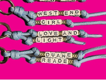May include: Keychains with white letter beads spelling out phrases like "WHAT A SAD MAN" and "WEST END GIRL." The keychains have blue cords and gold-colored accents, set against a bright pink background. Each keychain has a gold-colored ring.