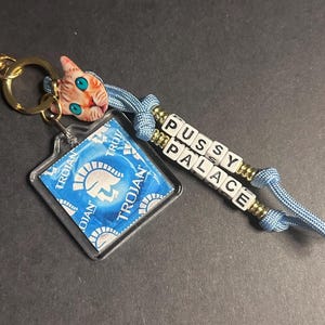 May include: A novelty keychain featuring a gold-colored clasp, a clear square with a blue Trojan condom, a cat-shaped charm, and a beaded strand spelling "PUSSY PALACE". The keychain has blue cord accents.