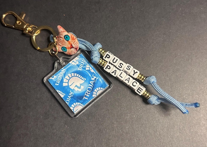 May include: A novelty keychain featuring a gold-colored clasp, a clear square with a blue Trojan condom, a cat-shaped charm, and a beaded strand spelling "PUSSY PALACE". The keychain has blue cord accents.