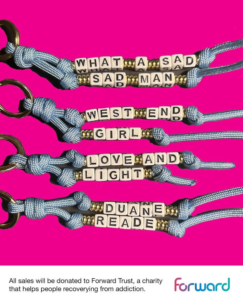 May include: Keychains with white letter beads spelling out phrases like "WHAT A SAD MAN" and "WEST END GIRL." The keychains have blue cords and gold-colored accents, set against a bright pink background. Each keychain has a gold-colored ring.