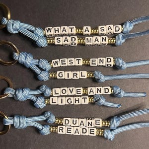 May include: Four handmade keychains with light blue cord and gold-colored metal rings. Each keychain features white letter beads spelling out phrases: "WHAT A SAD MAN", "WEST END GIRL", "LOVE AND LIGHT", and "DUANE READE".