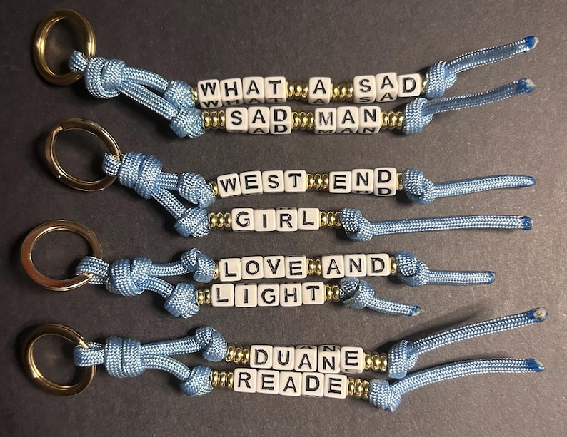 May include: Four handmade keychains with light blue cord and gold-colored metal rings. Each keychain features white letter beads spelling out phrases: "WHAT A SAD MAN", "WEST END GIRL", "LOVE AND LIGHT", and "DUANE READE".