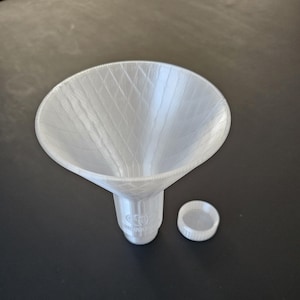 Könnte beinhalten: Ein weißer Kunststofftrichter mit einem breiten, konisch geformten Oberteil und einem schmalen Ausguss. Ein kleiner, runder Deckel liegt neben dem Trichter. Trichter und Deckel bestehen aus einem strukturierten, 3D-gedruckten Material.