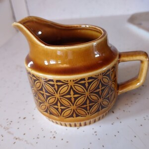 May include: A brown ceramic creamer with a geometric pattern in brown and black. The creamer has a rounded body and a handle.