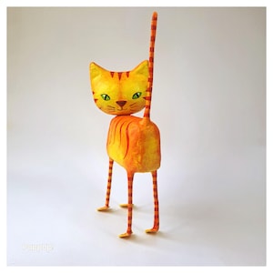 Könnte beinhalten: Eine skurrile, handgefertigte Katzenfigur in Gelb und Orange. Die Katze hat einen großen Kopf, grüne Augen und einen gestreiften Schwanz. Der Körper ist orange mit vertikalen Streifen. Sie steht auf dünnen, gestreiften Beinen. Eine einzigartige, künstlerische Katzenfigur.
