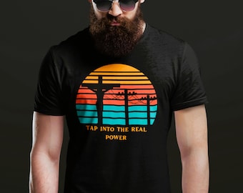 May include: A black t-shirt featuring a graphic of a sunset with horizontal stripes in orange, yellow, and teal. The design includes a silhouette of a cross and utility poles with power lines. The text reads, TAP INTO THE REAL POWER.