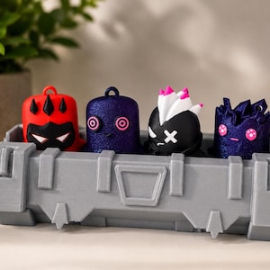 May include: Four small, stylized character figurines with loops on top, displayed inside a rectangular gray storage crate. The figures include a red one with black spikes, a glittery purple one with spiral eyes, a black one with white hair, and a dark purple one.