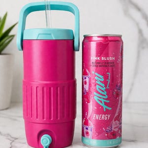 Op de afbeelding: Een roze geïsoleerde waterkan met een turquoise handvat en deksel, naast een roze blikje Alani Energy Pink Slush-drank. Op het blikje staat de tekst "Pink Slush" en "Energy". De kan heeft een helder rietje.