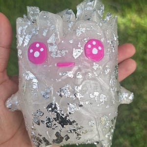 May include: A small, translucent, creature-shaped figurine made of resin with embedded silver foil flakes. The figure features two bright pink circular eyes with white highlights and a small pink horizontal line for a mouth, with jagged, crystal-like protrusions on the top.