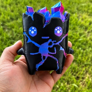 May include: A small, black, hollow figurine with a jagged top opening revealing bright blue and magenta crystalline structures inside. The front features two large, round, blue and magenta eyes and a central crack pattern in the same vibrant colors.