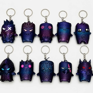 May include: A collection of ten small, character-themed keychains arranged in two rows. Each figure features a gradient finish shifting between deep blue and purple, with unique facial expressions, horns, hoods, or markings rendered in bright colors.