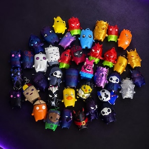 May include: A collection of small, colorful plastic character figurines featuring various designs, including skulls, animals, and abstract shapes in shades of red, blue, yellow, purple, orange, and black, arranged in a cluster on a dark surface.
