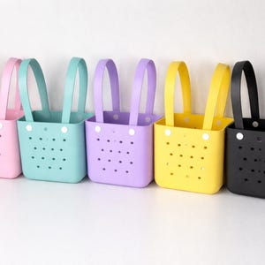 Puede incluir: Una fila de seis bolsas de mano coloridas en tonos pastel de rosa, turquesa, morado, amarillo y negro. Cada bolsa tiene un asa a juego y está hecha de un material perforado. Las bolsas están alineadas sobre un fondo blanco.