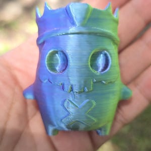May include: A small, 3D-printed figurine featuring a gradient of blue, purple, and green colors. The character has a crown on its head, large oval eyes, a jagged mouth line, and an X-shaped mark over its mouth area, with a smooth, layered texture.