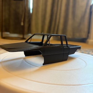 May include: A matte black model pickup truck, showcasing the cab and bed. The vehicle is positioned on a white surface, with the focus on the detailed design of the truck's body. The model appears to be a scale replica.