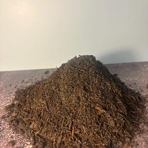 以下が含まれることがあります： 細かく砕いた木材と有機物で構成された、濃い茶色のマルチングの山が、質感のある表面に置かれています。 マルチングは湿っていて栄養が豊富に見え、ガーデニングや造園に適しています。 背景は無地のオフホワイトです。