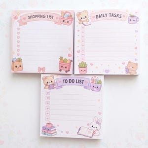 May include: Three square paper notepads with light pink and purple pastel themes. The top left pad says SHOPPING LIST, the top right says DAILY TASKS, and the bottom pad says TO DO LIST. Each features cute cartoon animals, food, and stationery icons.