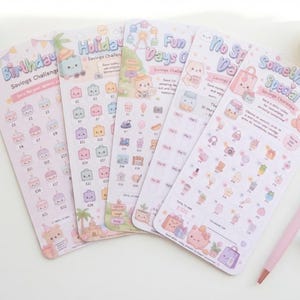 May include: Five rectangular paper savings challenge trackers with pastel illustrations and text including Birthday, Holiday, Fun Days, No Spend Days, and Something Special. Each tracker features rows of small icons with pound amounts for tracking.