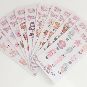 May include: A set of white rectangular savings challenge cards arranged in a fan shape. Each card features pastel illustrations of animals, mermaids, and food items, numbered sequences, and the text 100 Save £5,050 printed in pink script.