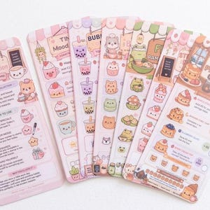 May include: A set of six rectangular paper cards featuring pastel-coloured illustrations of cafe-themed items, including bubble tea, ramen, and pancakes. Each card contains printed text, checkboxes, and tracking grids for daily habits and rewards.