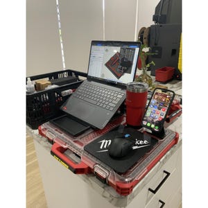 May include: A grey laptop sits on a red and clear plastic organiser case, displaying 3D modelling software. Next to it is a red insulated tumbler, a black computer mouse on a Milwaukee branded mousepad, and a smartphone in a black stand.