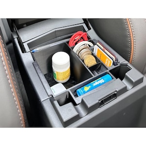 May include: A black plastic car centre console organiser insert with multiple compartments. It contains a white pill bottle, a blue Blistex lip balm, a stack of gold coins, a red charging cable, a clear pen, and several credit cards.