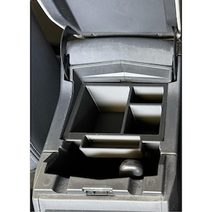 Op de afbeelding: Een zwarte kunststof auto middenconsole organizer inzetstuk met meerdere compartimenten van verschillende groottes. De lade is ontworpen om in de diepe opbergruimte van een voertuigconsole te passen voor het organiseren van kleine voorwerpen.
