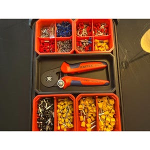 Puede incluir: Una bandeja organizadora de plástico negro que contiene una herramienta de crimpado Knipex con mangos naranjas y azules, rodeada de varios compartimentos rojos llenos de terminales y conectores eléctricos variados en rojo, azul, amarillo, gris y negro.